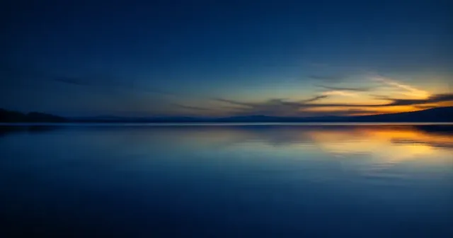 Un lago azul y tranquilo al anochecer, con una franja de luz naranja en el horizonte.