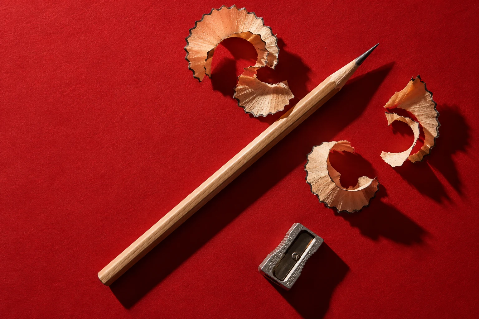 A sharply pointed wooden pencil and curled cedar shavings lie on a crimson surface.