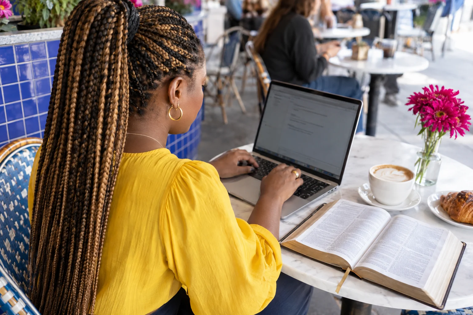 A woman with long braids types beside an open Bible in a cafe.