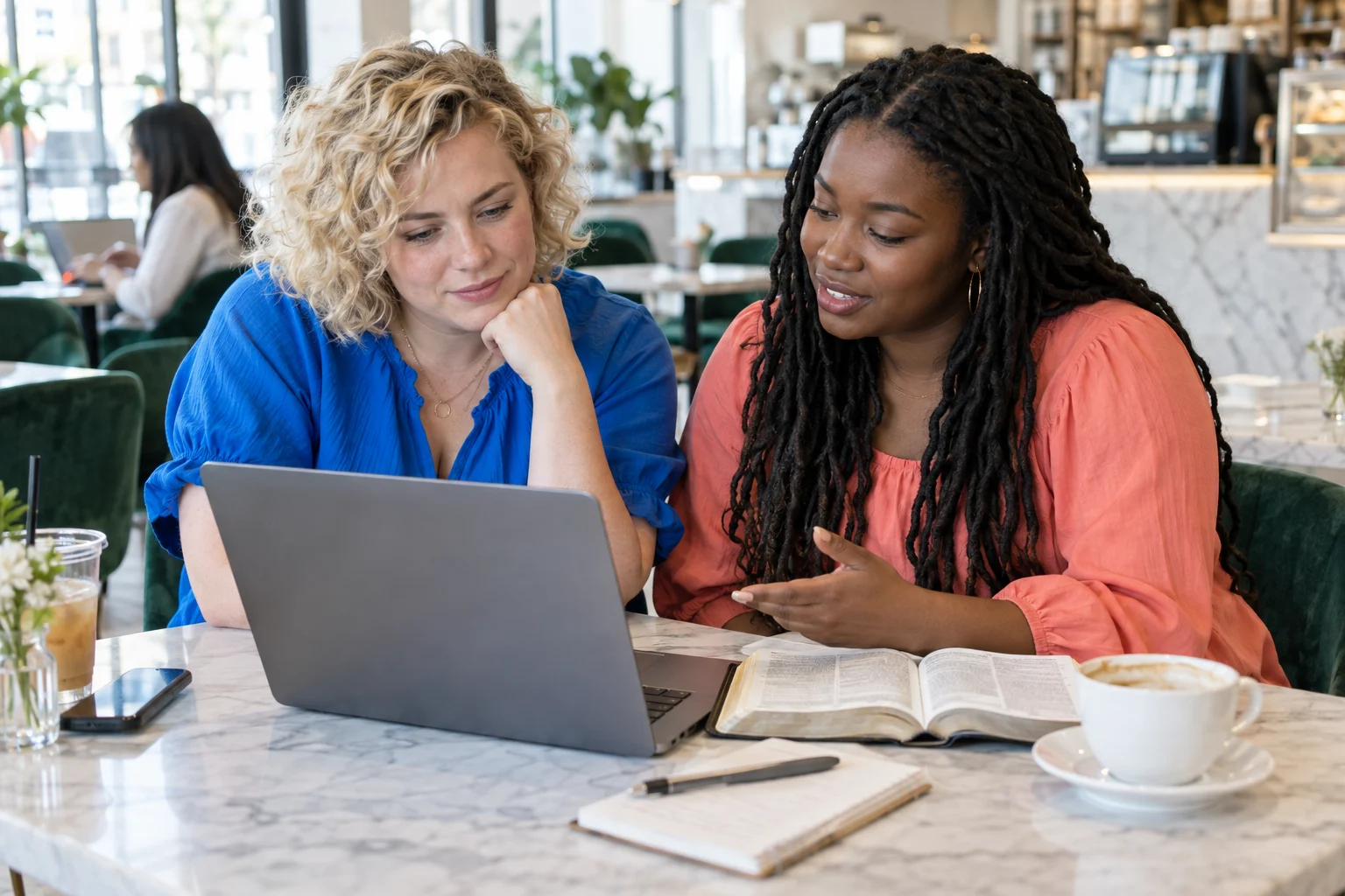 Two friends, one with blond curls and one with locs, study a Bible beside a laptop.